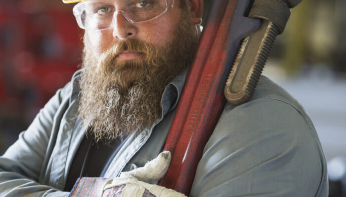 A bearded man working in a metal fabrication plant, carrying a large metal tool on his shoulder.